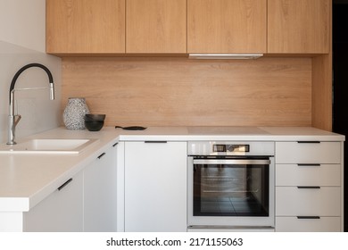 Simple kitchen with white cupboards, drawers and countertops, sink, oven and induction hob and wooden backsplash and top cabinets