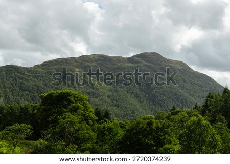 A simple landscape image of a tree covered hill rising towards a greay and cloudy sky in the Lake District. The steep sided mountain gives way to the dramatic highland fells.