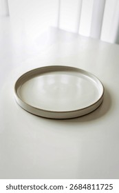 A simple, light gray ceramic plate with a thin, muted green edge rests on a bright white, slightly textured surface.