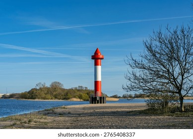A simple lighthouse and a leafless tree at the beach of Juliusplate on a sunny day in April with vivid blue sky - the Warflether Sand peninsula can be seen in the background