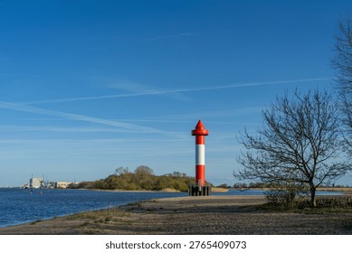 A simple lighthouse and a leafless tree at the beach of Juliusplate on a sunny day in April with vivid blue sky - the Warflether Sand peninsula can be seen in the background