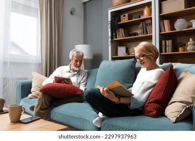 Simple living. Happy middle-aged senior couple 70s husband and wife sit relax on couch in living room reading book drinking tea together, calm elderly 60s couple booklovers rest at home enjoy life - Powered by Shutterstock