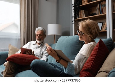 Simple living. Happy middle-aged senior couple 70s husband and wife sit relax on couch in living room reading book drinking tea together, calm elderly 60s couple booklovers rest at home enjoy life - Powered by Shutterstock