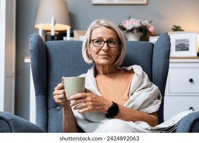 Simple living. Senior woman sitting alone on chair at home drinking tea or coffee enjoying her time and life. Older female thinking about her past and happiness, satisfied with her life decisions