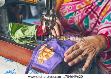 A simple looking Indian woman stitching handmade cloth face masks inside her home. Closeup of sewing machine needle, unfinished masks, cotton threads etc.