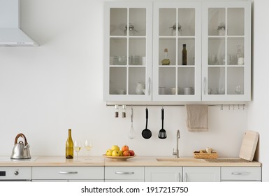 Simple modern kitchen, scandinavian design and new style after repair. Metal kettle on stove with hood. Colored fruits on plate, utensils, bottle of wine and glasses on white furniture and light wall