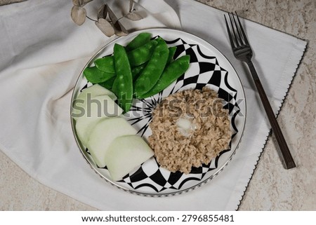 simple and nutricious breakfast featuring creamy oatmeal topped with a piece of butter, served with fresh green peas and kohlrabi on a modern white and black plate. Minimalist food styling with clean background, emphasizing healthy eating, wholesome ingredients and balanced nutrition. Ideal for healthy lifestyle, diet, wellness and breakfast concept imagery