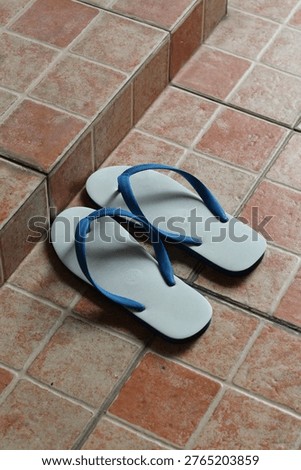 A simple pair of white flip-flops with bright blue straps are placed on a textured tiled surface, suggesting a casual or domestic setting