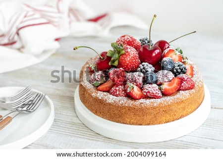 Simple round sponge cake with fresh summer fruits served on white wooden board; homemade sweet baked dessert topped with berries and cherries