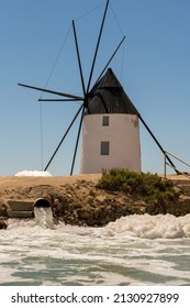 Simple Round White Windmill With A Brown Conical Roof. The Building, Typical Of Murcia, Is Next To A Wide Bore Pipe Pumping Salty Water Into A Dehydration Pond. Spume Fills The Foreground. Copy Space.