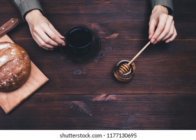 Simple rural breakfast with tea, honey and rye bread. Woman's hands holding spoon and cup on dark wooden table, top view. - Powered by Shutterstock