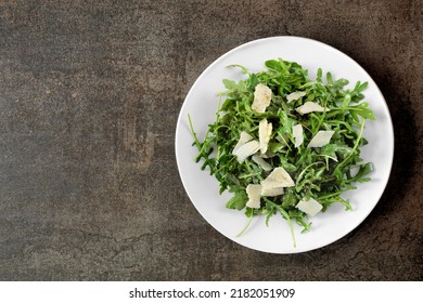 Simple Salad Of Arugula And Parmesan Cheese. Overhead View On A White Plate Over A Dark Background.