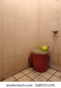 A simple, tiled bathroom corner featuring a red bucket with a green dipper, suggesting traditional or basic washing facilities. The light-colored tiles evoke a no-frills, functional space