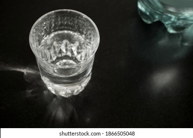 A Simple Water Glass With Water And A Part Of A Green Plastic Bottle On A Black Granite Slab From Top View Close-up