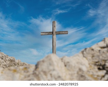 A simple wooden cross stands tall on a rocky hilltop against a backdrop of dramatic blue sky with clouds. - Powered by Shutterstock