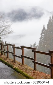 A simple wooden fence borders the cliffside, with trees and mountain partially visible through heavy fog and clouds in the background. Miyazaki, Japan. Travel and nature concept.