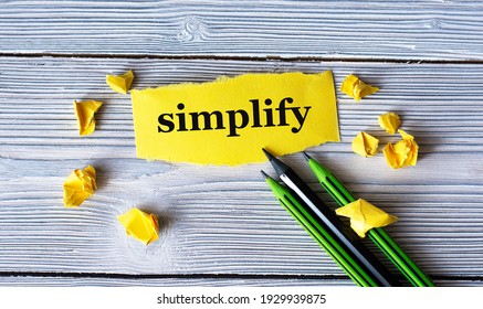 SIMPLIFY - word on a yellow tattered piece of paper with pencils and crumpled paper lumps on a light wooden background. Info concept