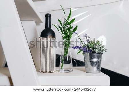 A simplistic home decor setting with a glass vase and fresh green plants beside a covered wine bottle on a white staircase.