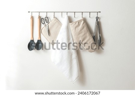 Simplistic Kitchen Wall Organization with Modern Utensils: Silicone-Tipped Wooden Spoons, Stainless Steel Scissors, Natural Fabric Potholder, White Terry Towel, and Tongs, all under Soft Lighting. Foto stock © 