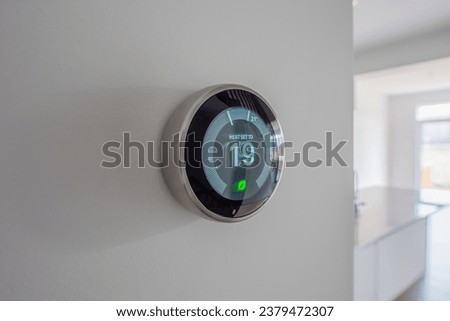 A simplistic photo of a round, modern, programmable digital thermostat, on a clean white wall in heating mode
