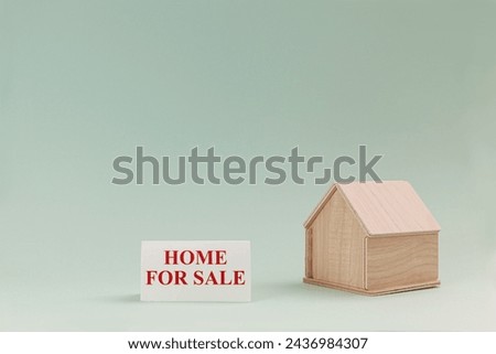 Simplistic wooden house model isolated on pale green background, with text Home For Sale on signboard.