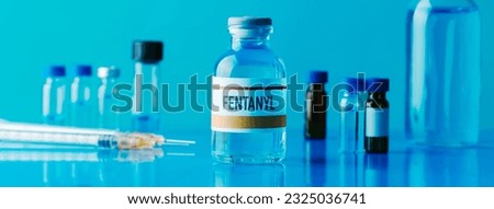 a simulated vial of fentanyl next to a syringe and some other different vials on a blue table, in a panoramic format to use as web banner