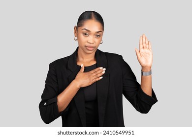 Sincere African American businesswoman making a pledge or swearing an oath with her right hand raised and left hand on her chest, in a professional black suit on grey background, studio - Powered by Shutterstock