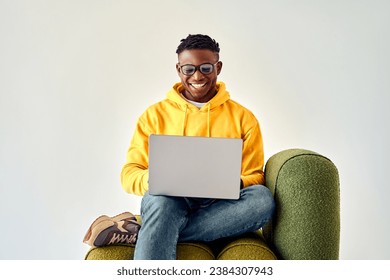 Sincere emotions and technology. Black male freelancer looking on laptop screen with cheerful smile while sitting on green lounge chair in studio. Young guy wearing googles and casual yellow hoodie. - Powered by Shutterstock