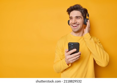 Sincere handsome man smiles gladfully wears headphones on ears listens favorite music uses cellphone dressed in casual jumper looks away being in good mood isolated over yellow studio background - Powered by Shutterstock