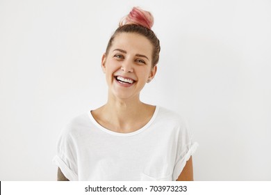 Sincere joy, happiness, youth and carefree life. Indoor shot of lovely hipster girl with hair bun dyed pink having fun, laughing out loud at some joke or funny story, dressed in stylish white t-shirt