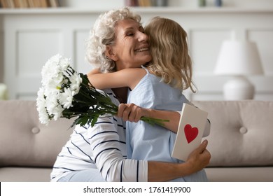 Sincere Old Grandma Hugs Small Attentive Granddaughter Express Her Gratitude For Received Surprise At Birthday Or 8-march Woman Day. Grandchild Makes Surprise For Granny Gave Her Flowers And Postcard