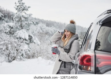 Sincere Smile. Pretty Girl In Warm Clothes Standing In The Winter Wood While Leans On The Car And Holding Cup Of Coffee.