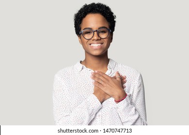 Sincere thankful smiling african american woman holding hands near heart on chest, looking at camera. Head shot close up portrait grateful woman in eyeglasses, isolated on grey studio background.