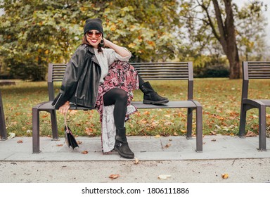 Sincerely laughing female dressed fashion boho style colorful long dress, warm knitted sweater with black leather biker jacket and Beanie Hat with flap bag sitting on bench at autumn city park - Powered by Shutterstock