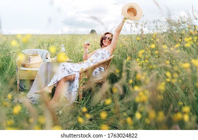 Sincerely smiling young Woman dressed  light summer dress sitting in rattan chair on the high green grass meadow with glass of red wine and cheerful rised arm up with straw hat.