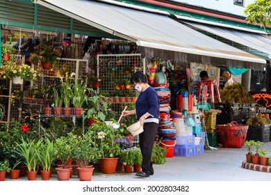 SINGAPORE - 5 JUL 2021: A Shop Owner Wears A White Reusable Face Mask With The Merlion Logo As She Waters Her Potted Plants Which Are  In High Demand As More People Work Remotely.