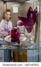 Singapore City, Singapore; January 23,2019: Dinner Setting Of Plate, Wine Glass, Silverware, And Folded Clothe Napkins On Display In Glass Case With Unidentified Lady In Background.