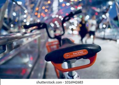 SINGAPORE - FEB 2, 2018: Grey And Orange Bicycle In Bike Sharing Project, Scan And Ride By Mobike Parking At City Street With Blurred Light Bokeh And Couple People At Night, Singapore