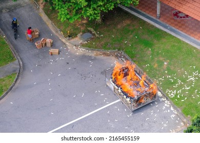 Singapore Sep2021 Large metal container of burning joss paper offerings by residents during Hungry Ghost Festival. Paper litter on tarmac ground. Smoke, ashes, dust specks in air