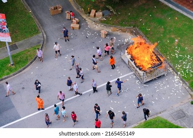 Singapore Sep2021 People burning throwing joss paper money into metal cage, offerings during 7th month Hungry Ghost Festival. Pollution with litter on tarmac, ashes from huge fire, dust specks in air