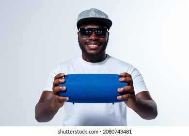 A singer in cap is showing a blue bluetooth speaker to us. His cap is gray. He wears dark glasses. He has a beard. He is holding a cool bluetooth speaker in his hands.