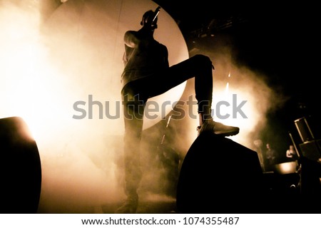 A Singer is on the stage. A silhouette of the singer is putting his foot on a speaker. A brutal shadow of a rapper on the stage. Smoke and bright soft yellow stage lights in the background.