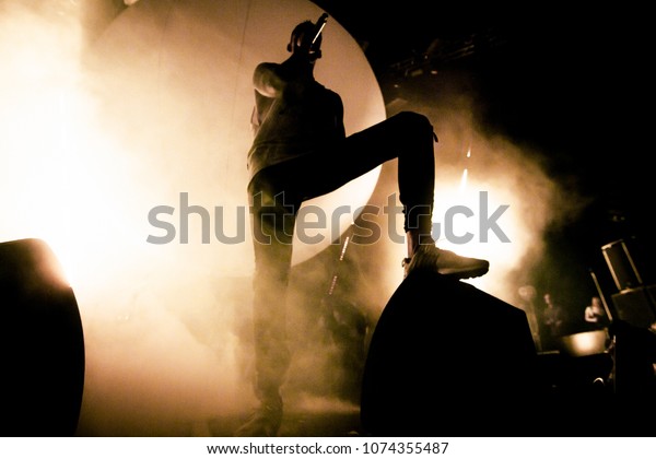 A\
Singer is on the stage. A silhouette of the singer is putting his\
foot on a speaker. A brutal shadow of a rapper on the stage. Smoke\
and bright soft yellow stage lights in the\
background.
