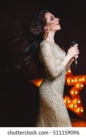 Singer Woman In Sexy Glitter Long Dress On Stage With Broadway Star On Background. Curly Hairstyle, Perfect Make-up. Action.