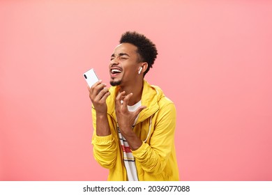 Singing from bottom of heart along favorite music listening song in wireless earbuds with closed eyes and joyful smile gesturing passionately making microphone from smartphone over pink background - Powered by Shutterstock