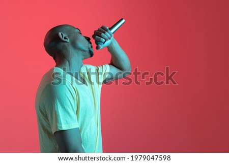 Singing, performing. African-american young man's portrait on red studio background in neon light. Beautiful male model in casual, rapping. Concept of human emotions, facial expression, youth, sales