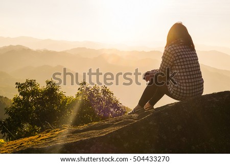 single adult woman silhouette on rock watching yellow and orange setting sun in distance