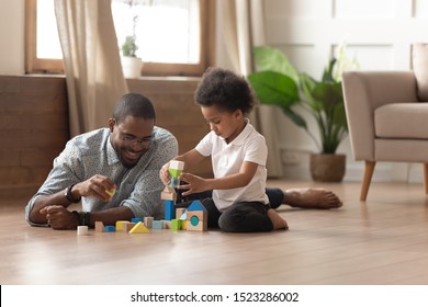 Single African American Smiling Father Or Male Babysitter Lying On Heated Warm Floor, Playing With Cute Little Boy, Constructing Tower From Bright Colorful Wooden Blocks, Leisure Family Activity.