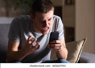 Single angry person with a mobile phone sitting on a sofa in the living room in a house indoor with a dark background