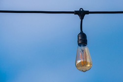 A Single Bare Outdoor Light Bulb Hangs From Its Socket On A Black Electrical Wire. The Isolated Bulb Is Part Of A String Of Lights And Is Suspended In Front Of A Blue Sky.